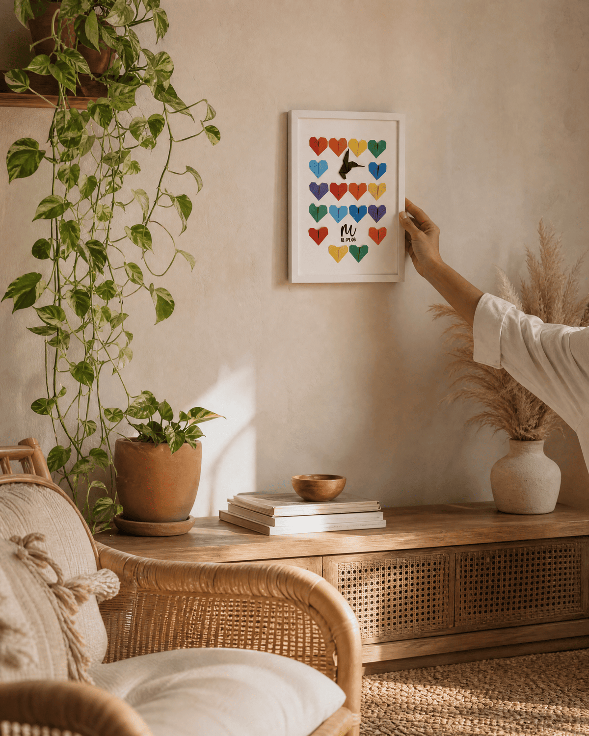 Mano colgando un cuadro Dos en Uno de corazones de origami de colores en una pared beige, sobre un aparador de ratán