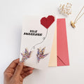 Tarjeta Feliz Aniversario de origami hecha a mano con corazón rojo y dos grullas de papel floral multicolor, sostenida en la mano junto a sus sobres