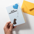 Tarjeta Feliz Cumpleaños de origami hecha a mano con corazón azul y perrito de papel marrón, sostenida en la mano junto a un sobre amarillo