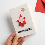 Tarjeta Feliz Navidad de origami hecha a mano con Papá Noel rojo y estrella dorada, sostenida en la mano junto a un sobre rojo
